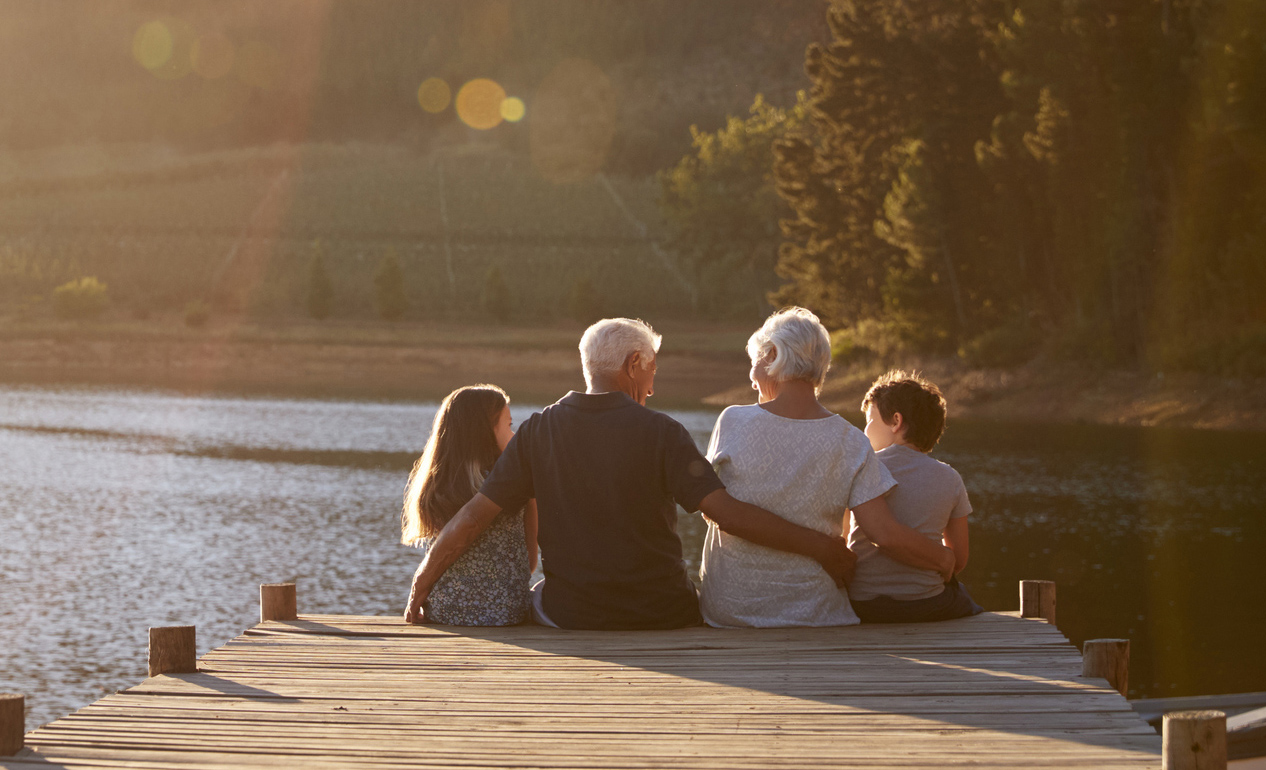 Grandchildren With Grandparents Sitting On Wooden Jetty By Lake